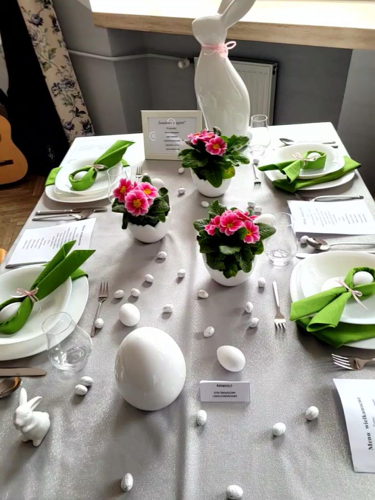 Zdjęcie - Prostokątny stół nakryty biało srebrzystym obrusem. Na obrusie znajduje się biała zastawa dla 4 osób. W każdym głębokim talerzu ułożona jest zielona serwetka złożona w uszy królika, w środku serwetki umieszczone jest białe jajko.
