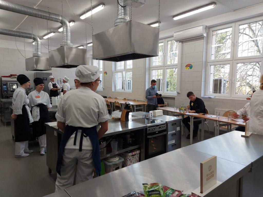 6 uczestników konkursu kulinarnego i trzech jurorów podczas konkursu w sali technologicznej, uczestnicy wykonują dania, jurorzy oceniają