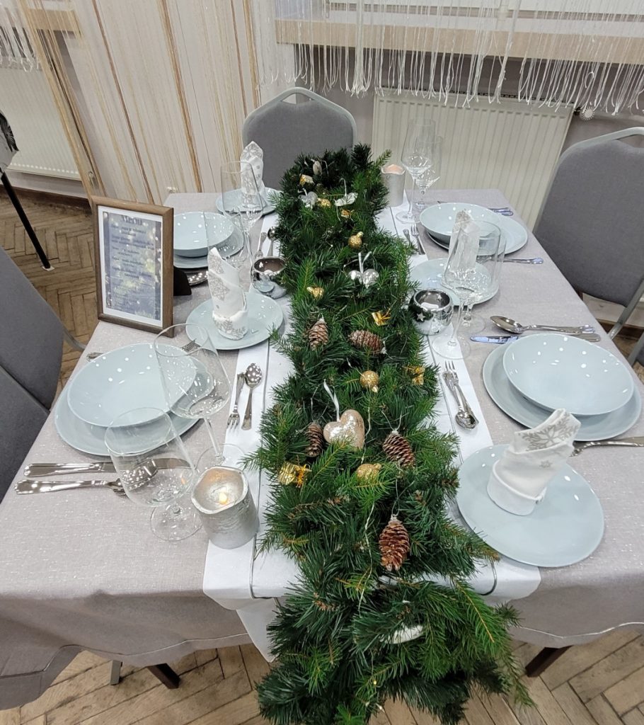 Zdjęcie - Obiad Bożonarodzeniowy  Jasny obrus, biały bieżnik na nim ułożony świerk z delikatną dekoracją i lampkami