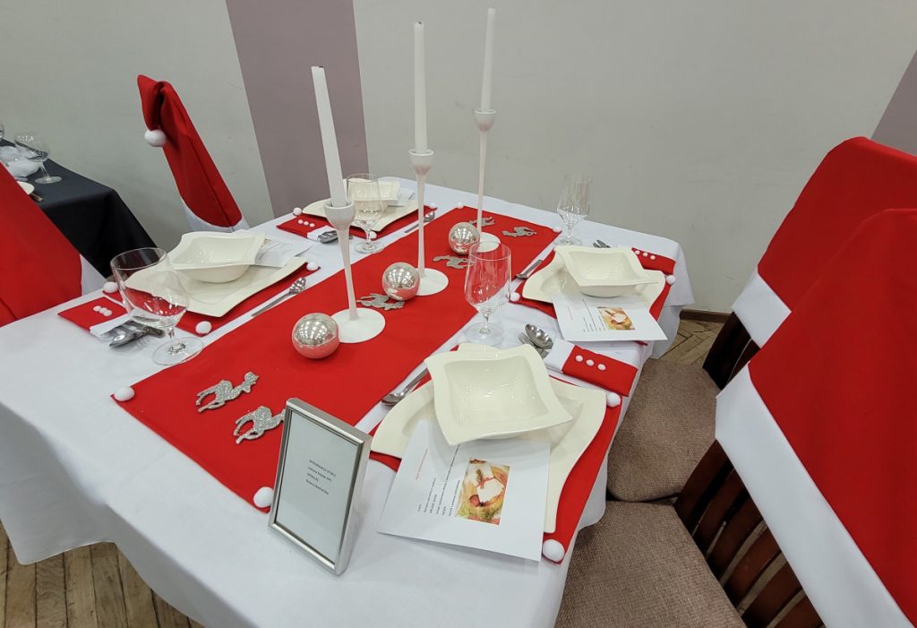 Zdjęcie - Wigilia biało-czerwona Biały obrus, czerwony bieżnik i czerwone podkładki pod białe talerze, po środku stołu ustawione wysokie świeczniki z białymi świeczkami, na krzesła nałożone są pokrowce czerwone z białymi pomponami