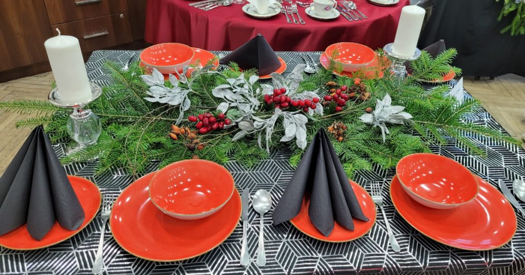 Zdjęcie - Stół zimowy,  Obrus czarno- srebrny w geometryczne wzory, czerwona zastaw stołowa, czarne serwetki, po środku ułożony świerk ze srebrno - czerwoną dekoracją