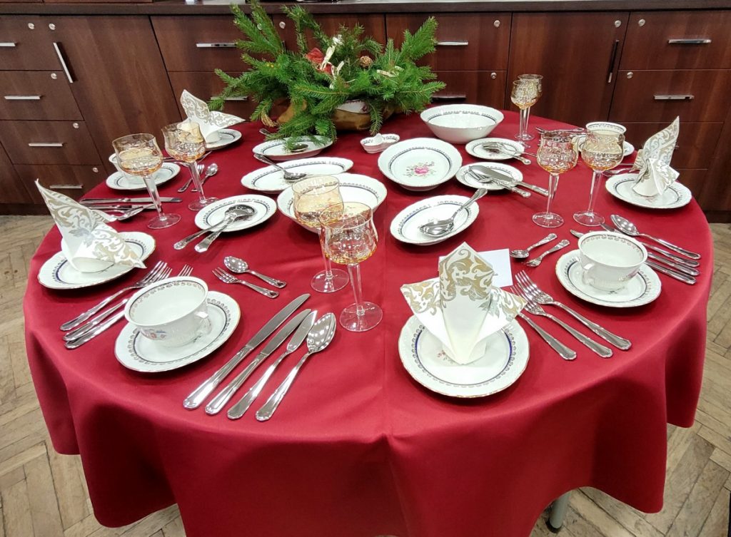 Zdjęcie - Tradycyjna Wigilia Okrągły stół nakryty bordowym obrusem, zastawa stołowa z motywem kwiatowym, herbaciane kieliszki, białe serwetki z motywem srebrno złoty, dekoracja ze świerku