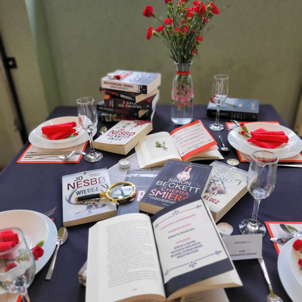 Zdjęcie - Wyróżnienie-wieczór z kryminałem. Czarny obrus białe talerze na nich  czerwone serwetki  na kształt uformowane w rozłożonej książki. Po środku wyeksponowano książki ”kryminały”.
Na końcu stołu ustawiony szklany wazon z czerwonymi goździkami.
