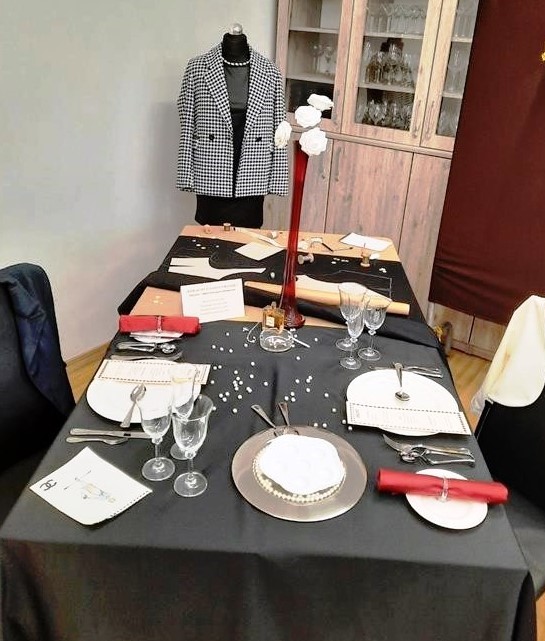 Zdjęcie - Stół prostokątny przygotowany dla dwóch osób nakryty do połowy czarnym obrusem. Na obrusie umieszczona biała zastawa stołowa. Miedzy nakryciami dla gości na srebrnym pod talerzu umieszczono półmisek do ślimaków. 