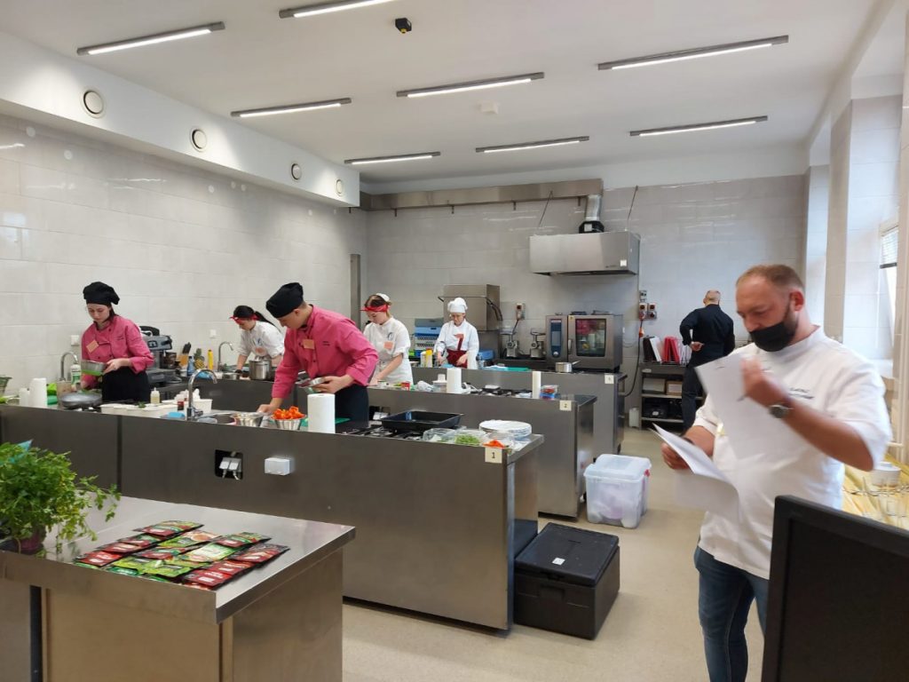 5 uczestników konkursu kulinarnego i dwóch jurorów podczas konkursu w sali technologicznej, uczestnicy wykonują dania, jurorzy oceniają