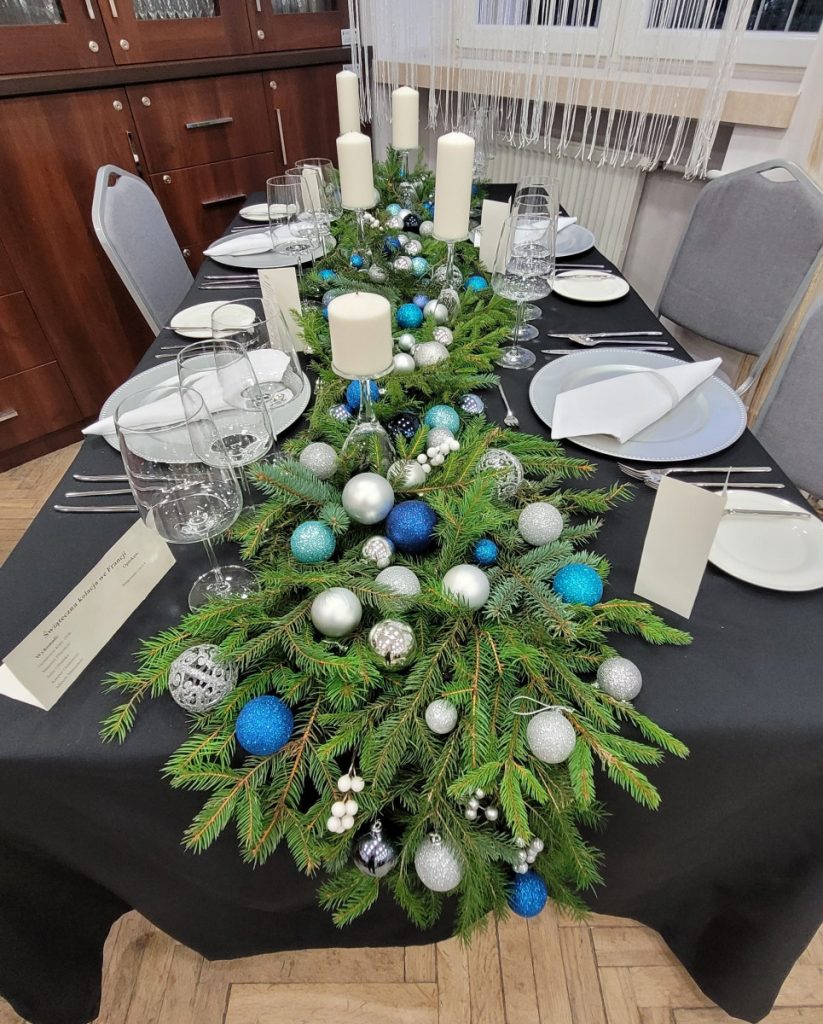 zdjęcie - 1.	Świąteczna kolacja we Francji Grafitowy obrus, biała zastawa stołowa, po środku ułożony świerk na nim bombki biało - niebiesko - granatowe