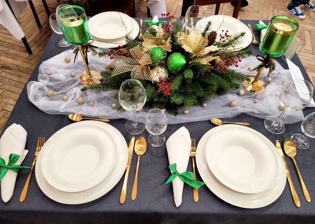 Zdjęcie - Stół Bożonarodzeniowy Szary obrus , białe serwetki i talerze, złote sztućce, zielone świeczniki, po środku stroik ze świerku z dekoracją zielono-złoto- czerwoną