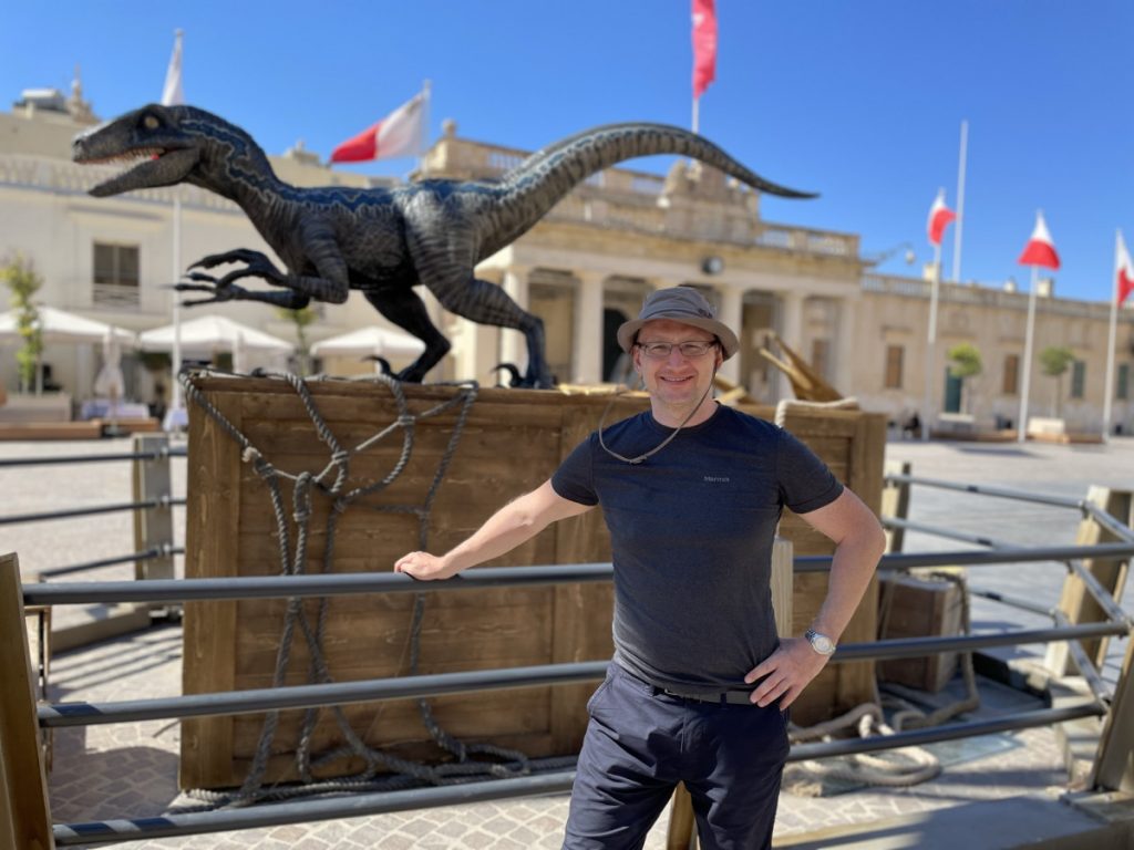 Przemysław Otto na St. George’s Square w Valetta przed skrzynią z dinozaurem z filmu Jurassic World Dominion