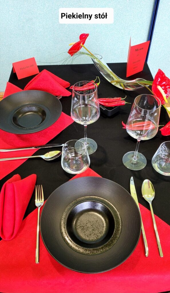 Zdjęcie - Na czarnym obrusie ułożone czerwono-bordowe serwetki a na nich czarne głębokie talerze. Dekoracja są papryczki czerwone chili ułożone w czarnych pojemniczkach, i czerwone anturium w kieliszku w kształcie cygara.